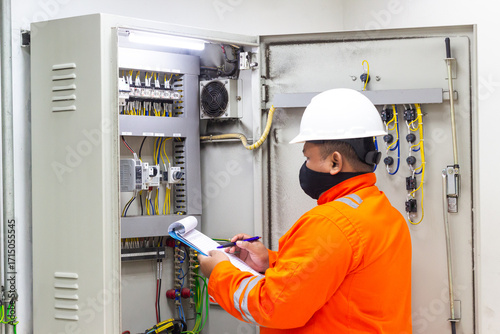 Electrical Engineer Inspecting Control Panel Wiring and Components, wearing hard hat and face mask, performing maintenance check while holding clipboard, ensuring safety and reliability