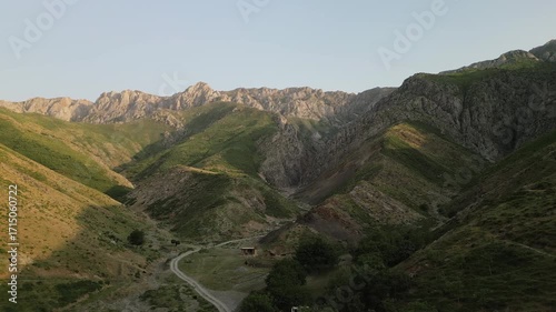 Aerial view down a narrow valley with a riverbed and farm clearing in Baysuntau, Surkhandarya. The drone tracks a gravel road between steep slopes, ideal for tourism, trekking and nature