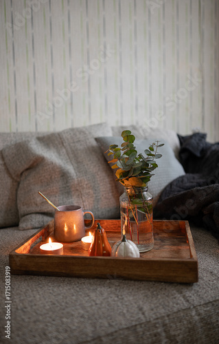 Cozy composition with cup of tea on the wooden tray, interesting book, candles and warm blanket. Hygge home interior.
Vertical image.