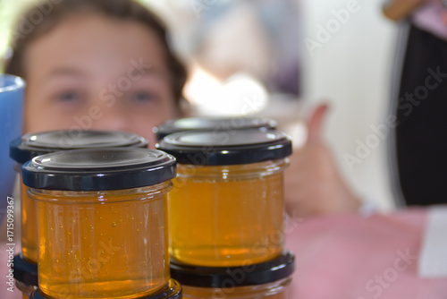Freshly filled glass jars of golden honey with black lids, stacked on a table. A child in the background smiles and gives a thumbs up, showing joy and approval