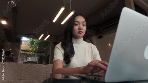 Confident young asian woman typing on a laptop computer while sitting in a cafe. Freelancer working remotely from a modern coffee shop