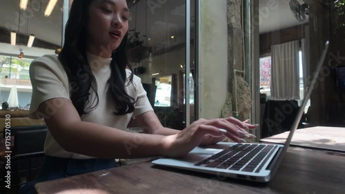 Young professional woman working on her portable computer, sitting at a wooden table in a sunlit coffee shop. Digital nomad lifestyle concept