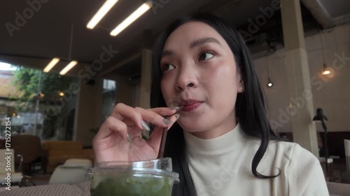 Stunning young Asian lady enjoying a chilled matcha latte while sitting in a trendy cafe, happily stirring and sipping her drink