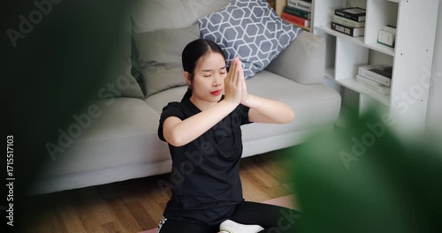 Handheld high angle view shot, Calm Asian woman is enjoying yoga in lotus position on a yoga mat in living room at home. Healthy Lifestyle Concepts