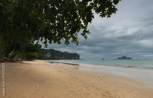 Scenic view of Ao Nang Beach in Krabi, Thailand, with golden sand, overhanging tropical trees, calm sea, and dramatic limestone cliffs under a cloudy sky.