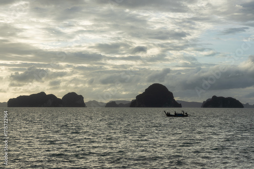 Traditional Thai fishing boat sailing on the Andaman Sea off the coast of Krabi, Thailand, with dramatic limestone islands silhouetted against a cloudy sky at dusk.