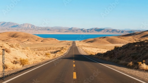 Straight road leading to a lake surrounded by desert hills and mountains under a clear blue sky