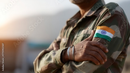 Proud Indian adult soldier wearing camouflage uniform with national flag patch on sleeve, standing with arms crossed in outdoor military setting, representing defense service with blurred background