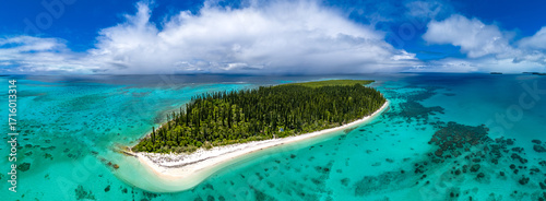 A wide aerial panorama of the remote and uninhabited Ilot Brosse in New Caledonia. The curved, pine-covered island is surrounded by a spectacular turquoise coral reef