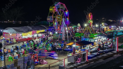 Bustling night market carnival illuminated by vivid, multicolored Ferris wheels. Large crowd people gather, enjoy various food stalls offering diverse culinary delights. Amusement rides attract