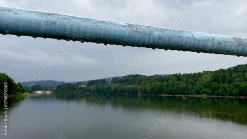 Close up video of a droplet falling from a metal railing with blurred lake and forest in the background.