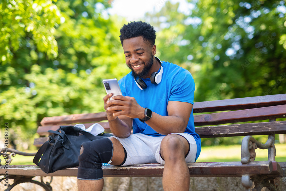 Fototapeta premium Young man sitting on bench with headphones and smartphone smiling outdoors after exercise. 