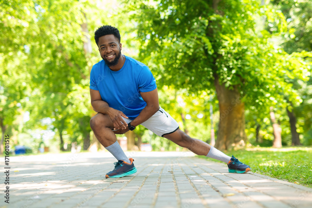 Obraz premium Smiling athlete stretching legs before outdoor workout. Young man in sportswear warming up in park on a sunny day.