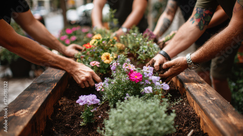 Hands planting flowers in wooden garden box with vibrant blooms against urban backdrop in sunny weather with greenery and soil involvement in community gardening activity