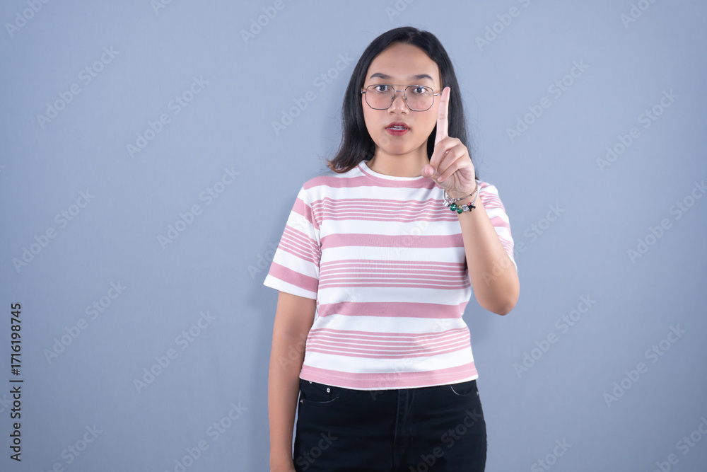 Fototapeta premium Young Asian woman in striped shirt and glasses, pointing finger upwards, serious expression