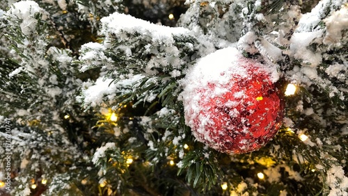 Snowy Christmas tree with red ball and lights garland. Urban city decoration at street. Xmas holiday. Close up. Winter. Outdoors.