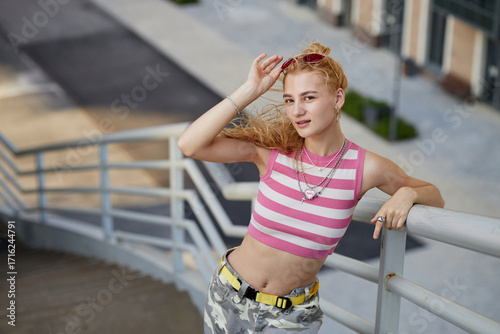 Portrait of Caucasian female zoomer standing outdoors on staircase holding sunglasses and smiling at camera, wearing layered necklaces and casual streetwear, urban background visible