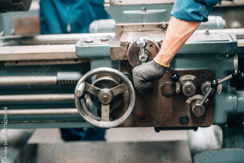 Slika na platnu A close-up shot shows a factory worker's hand, wearing a gray knit glove with a yellow cuff, operating a large industrial lathe
