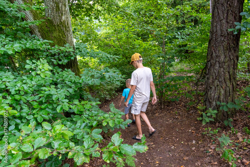 Two children walking together on forest trail surrounded by lush green vegetation and tall trees during summer nature hike. Family outdoor activities, childhood exploration and wilderness adventure.