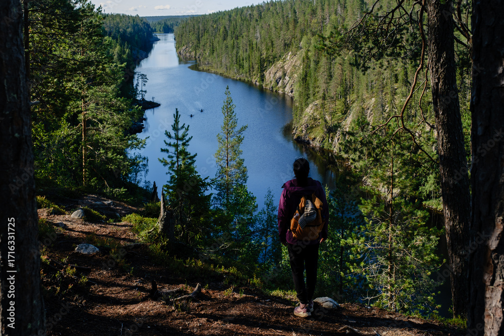 Fototapeta premium A solitary hiker stands overlooking the serene Canyon Lake Julma Olkky, surrounded by lush forest. The vibrant blue waters reflect the lush greenery, showcasing natures peace in Hossa National Park.
