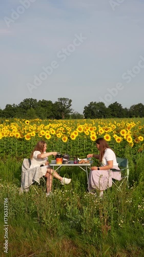 Two women enjoying outdoor picnic at small table in blooming sunflower field during summer day with thermos and snacks. Concept of female friendship, nature dining and countryside relaxation. 