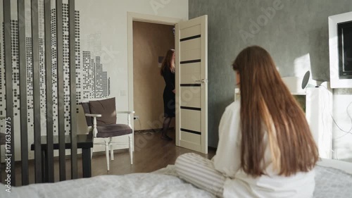 Students in modern bedroom, one girl exits through open door while friend sits on bed in striped pajamas watching, chairs, pillow, iron visible, casual dorm morning scene with calm mood