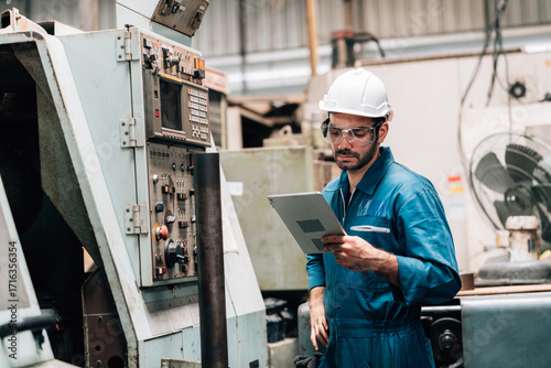 A young male factory worker, wearing a blue jumpsuit, a white hard hat, and safety glasses, is standing in front of a complex piece of industrial machinery. He is holding and looking down at a tablet