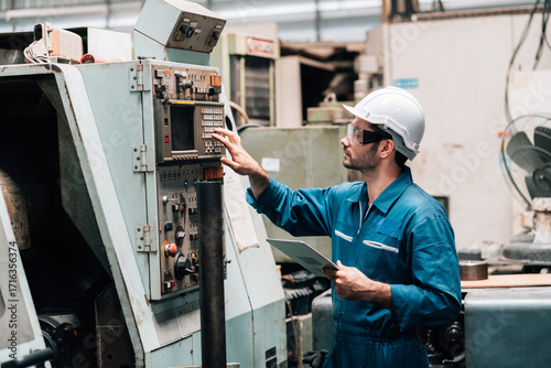 A young male factory worker, wearing a blue jumpsuit, a white hard hat, and safety glasses, is standing in front of a complex piece of industrial machinery. He is holding and looking down at a tablet