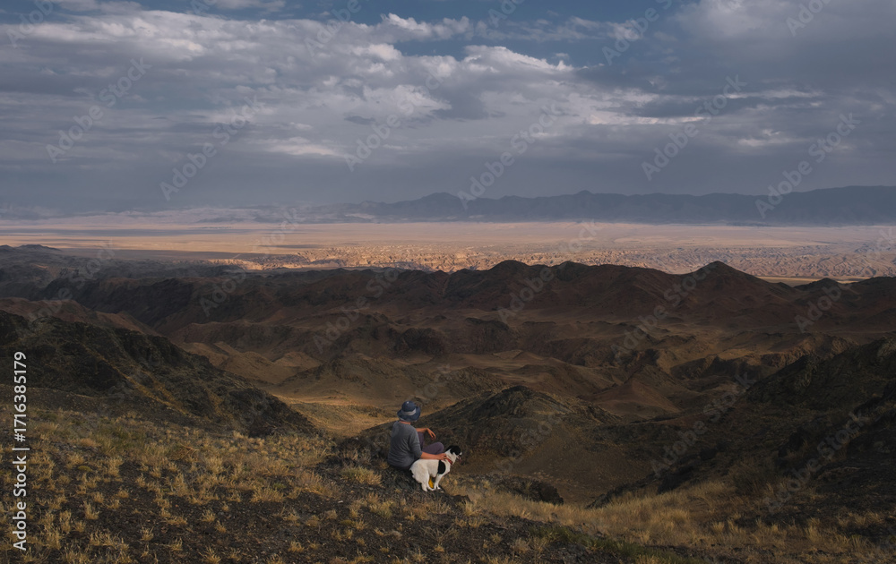 Obraz premium A woman in a hat sits with her dog on her back on a hill above the rugged desert mountains near the Charyn Canyon in Kazakhstan.