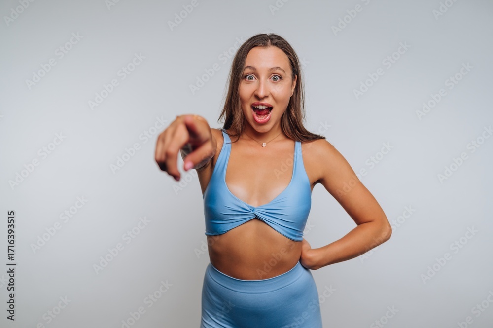 Fototapeta premium Woman in light blue activewear celebrates with excitement in a studio setting while pointing towards the camera