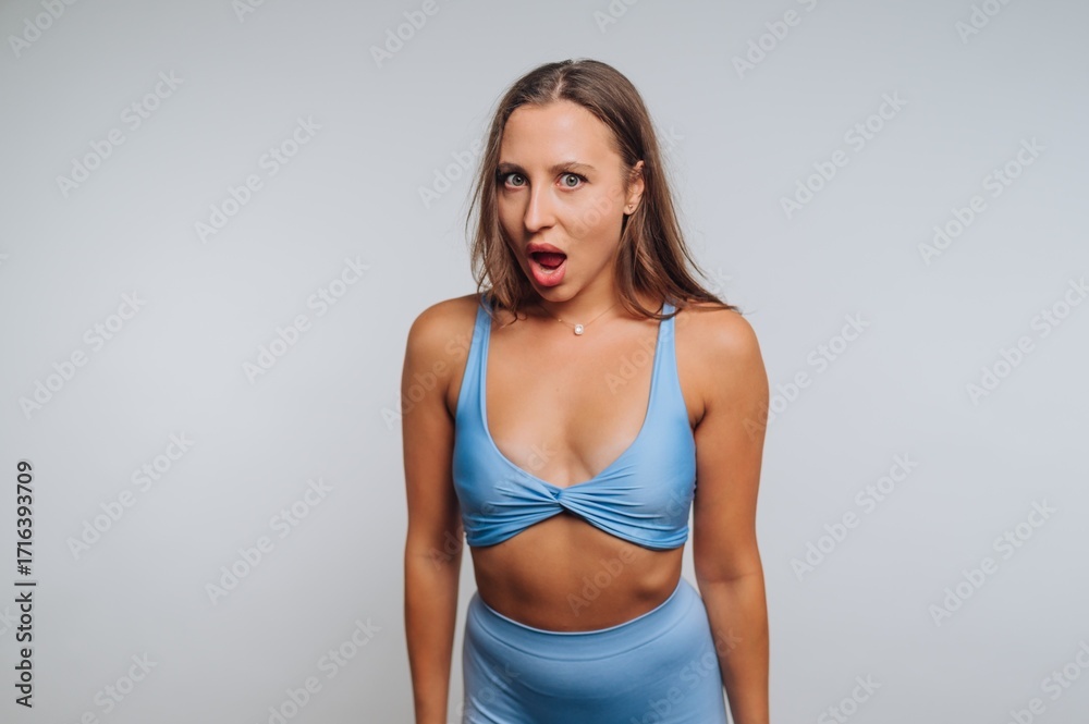 Fototapeta premium Young woman expressing surprise in stylish blue activewear during a studio photoshoot in a minimalistic setting