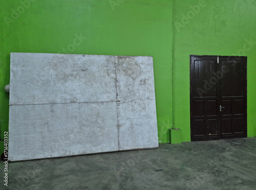 A large, weathered board leaning against a bright green wall next to a closed, dark wooden door on a concrete floor.
