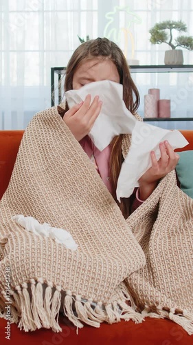 Ill girl child suffering from cold flu allergy infection wrapped in blanket sitting on sofa in living room at home. Sick teenager blows sneezes wipes snot into napkin. Coronavirus quarantine pandemic