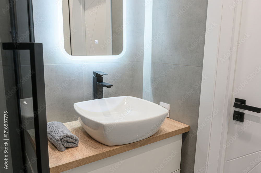Fototapeta premium Modern bathroom with a white square sink, black faucet, and illuminated mirror on gray tiles. Neutral tones and sleek design create a clean aesthetic
