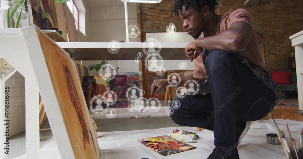 Fototapeta premium Crouching artist painting canvas on studio floor, with brush, art supplies and user icons overlay