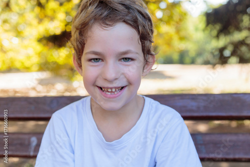 A close-up portrait of a happy, smiling boy with curly hair and sparkling blue eyes, sitting on a bench with a warm, autumn-themed background