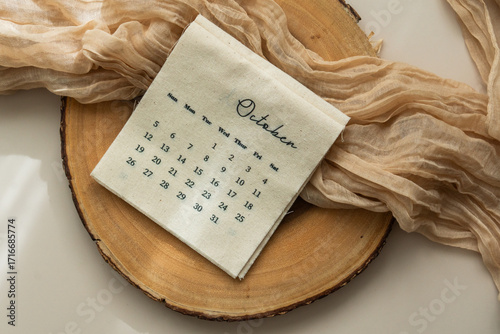 Top view of linen fabric calendar with October month, sheer fabric, wooden slice, light background, copy space, aesthetic soft shadows
