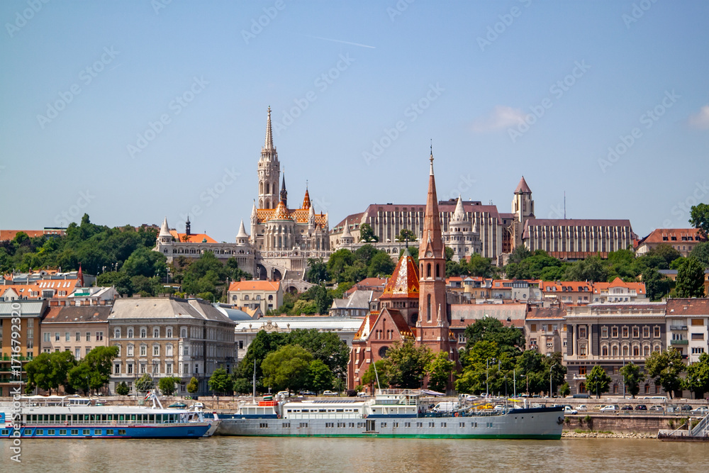 Fototapeta premium Panoramic view of vibrant Budapest cityscape along Danube River, showcasing iconic Matthias Church, historic spires, and diverse architecture, with multiple boats docked on sunny day.