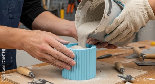 Two people are pouring a white liquid into a blue ribbed mold on a wooden table, with tools scattered around.