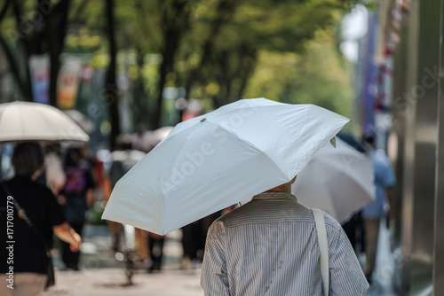 Man using sun umbrella for shade shows small sweat spots on shirt back, as he walks along tree lined sidewalk dotted with other pedestrians similarly shielding from midday sun heat in urban setting.