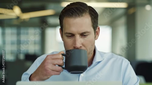 Man in blue shirt drinking from a dark mug while looking at a laptop in an office setting indoor