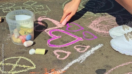 A child's hand draws hearts on the asphalt with pink chalk on a sunny day. A bucket with colorful chalk is nearby. A scene of creativity and children's outdoor games.