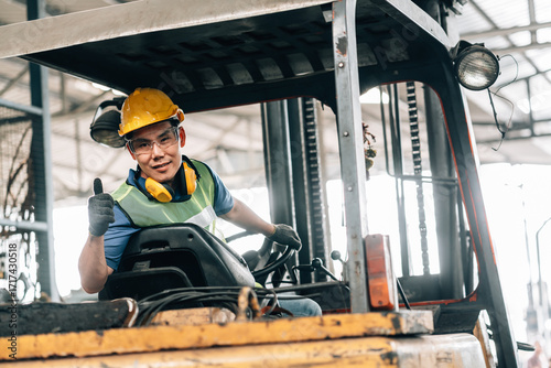 young male asian worker is sitting in the driver's seat of a forklift in a warehouse. The worker is looking directly at the camera with a confident smile and is giving a thumbs-up