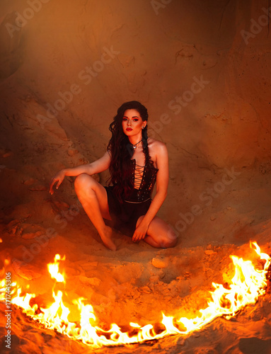 A young woman dressed in leather sits on the sand and looks through the flames of the fire in front of her.