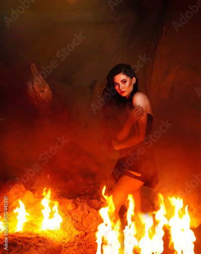 A young woman dressed in leather stands on the sand and looks through the flames of a fire in front of her.