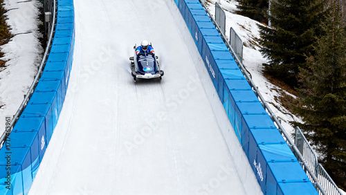 Athletes competing in a bobsled race on an icy track surrounded by blue barriers and snow-covered trees.