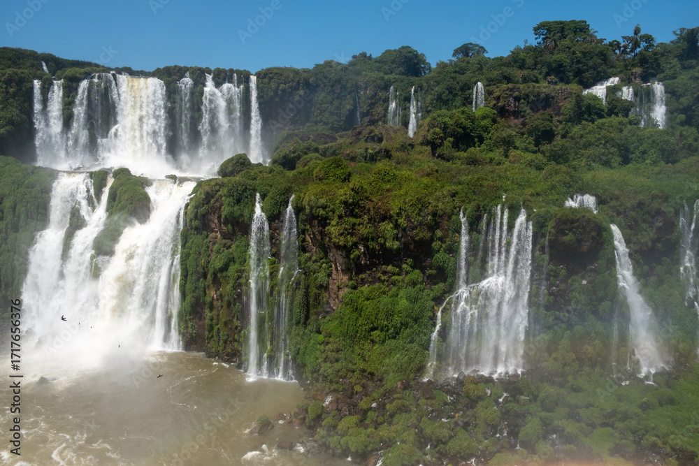 Fototapeta premium Iguazu Falls, showcasing its multiple, powerful cascades and a vibrant rainbow arcing through the mist. The shot captures the vast scale and stunning natural beauty of the site, making it ideal for to