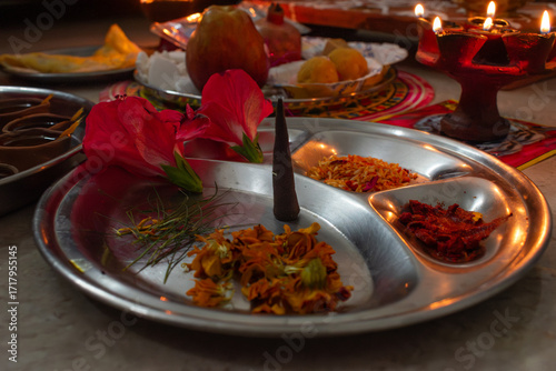 A traditional Hindu pooja thali with flowers, incense cone, turmeric, kumkum, and offerings arranged for a religious ceremony. Diya lit up in background.
