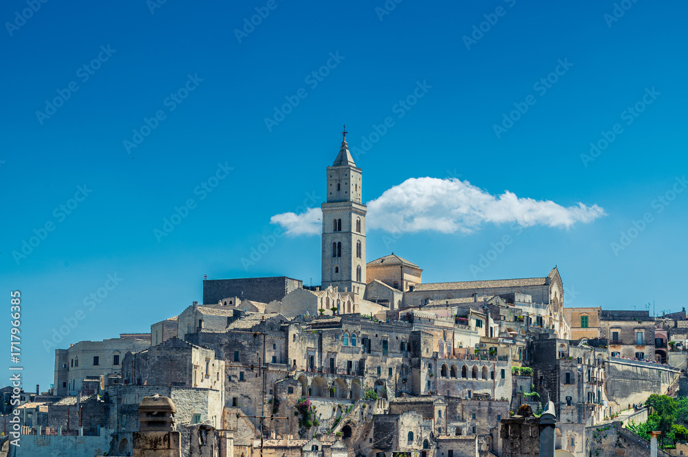 Obraz premium Vue du clocher de la Cathédrale de Matera et son quartier environnant sur fond de ciel bleu, avec un nuage derrière le clocher, Italie