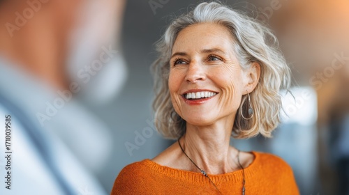 Smiling older woman with gray hair looks at a companion, wearing an orange sweater, exuding warmth and happiness indoors